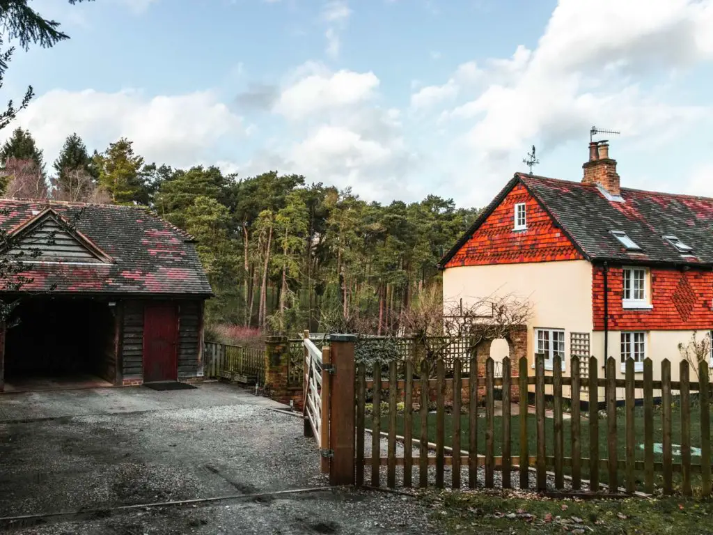 A cream and red house behind a fence, with a shed on the left, with a backdrop of trees. The sky is blue with white clouds.