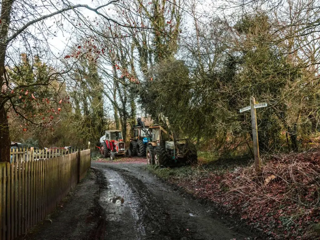 A first trail with a fence on the left and tractors parked up ahead. There is North Downs Way signage of the right.