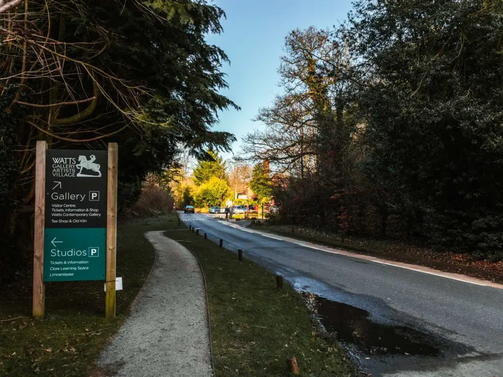 A road with bushes and trees either side. The road leads to a car park on the other end. There is signage for a gallery on the left. 