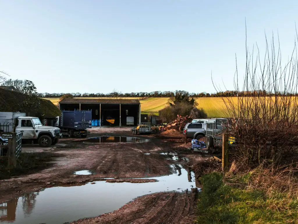 The shed and vehicles belonging to a farm. There is a view of a field in the background. The sky is blue.