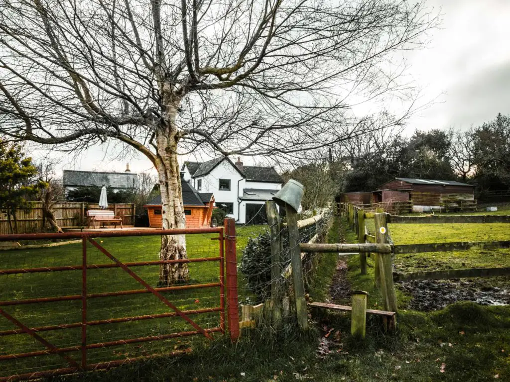 A fence leading to a grassy, muddy trail. There is a bucket resting g on the fence. There are houses in the background.