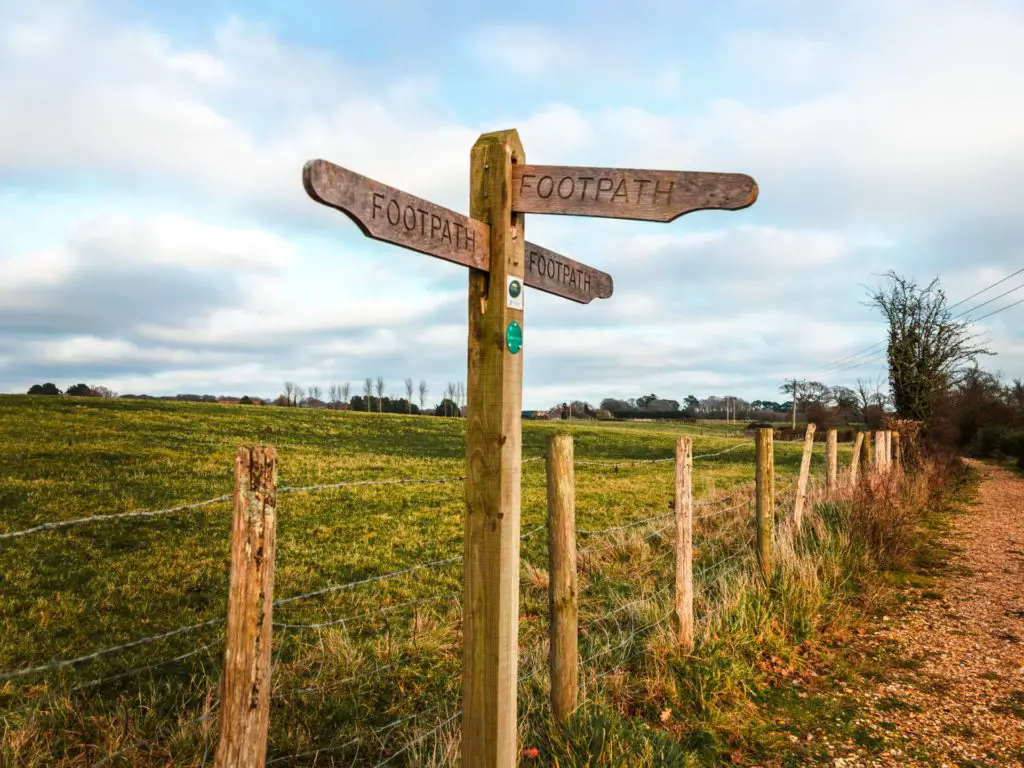 Walking signage on the trail in front of a large green field along the Brockenhurst to Lymington walk. The sky is blue with a few white clouds.