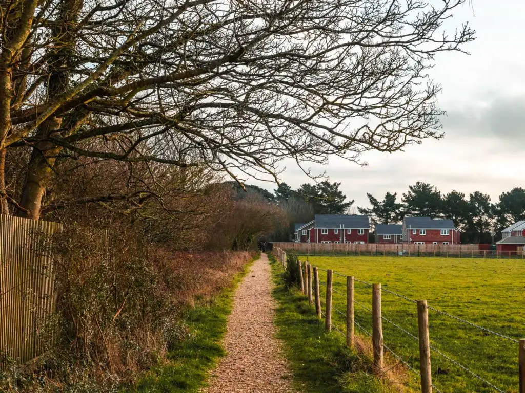 A gravel walking trail towards the end of the walk from Brockenhurst to Lymington. There is a barbed wire fence and green field on the right of the trail, and a fence and trees on the left. There are a few houses on the other end of the trail.