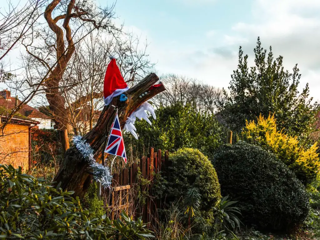 A tree stump decorated with a red Christmas hat, silver tinsel and the British flag. It is surrounded by green bushes.