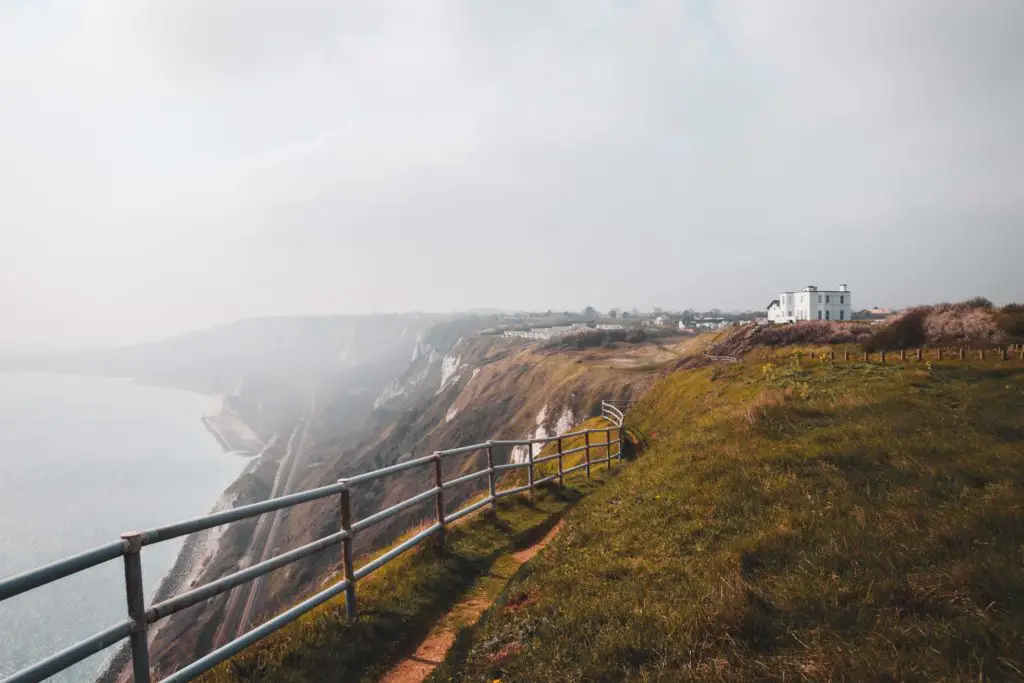 England Coast Path view when hiking from Folkestone to Dover on a misty day. The view is on top of the cliff with railings and a view of the ocean and train track down below. It's a misty day.