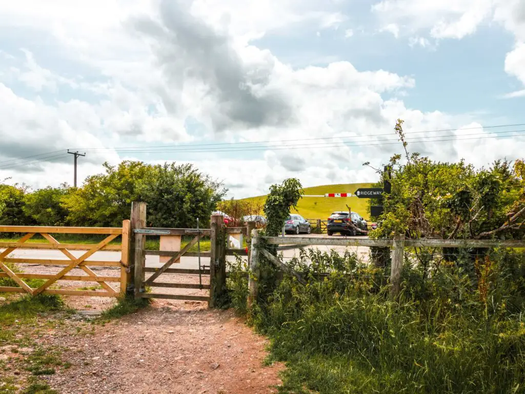 The trail as it leads to a gate and the main road. There are cars in the car park on the other side with a green hill in the distance.