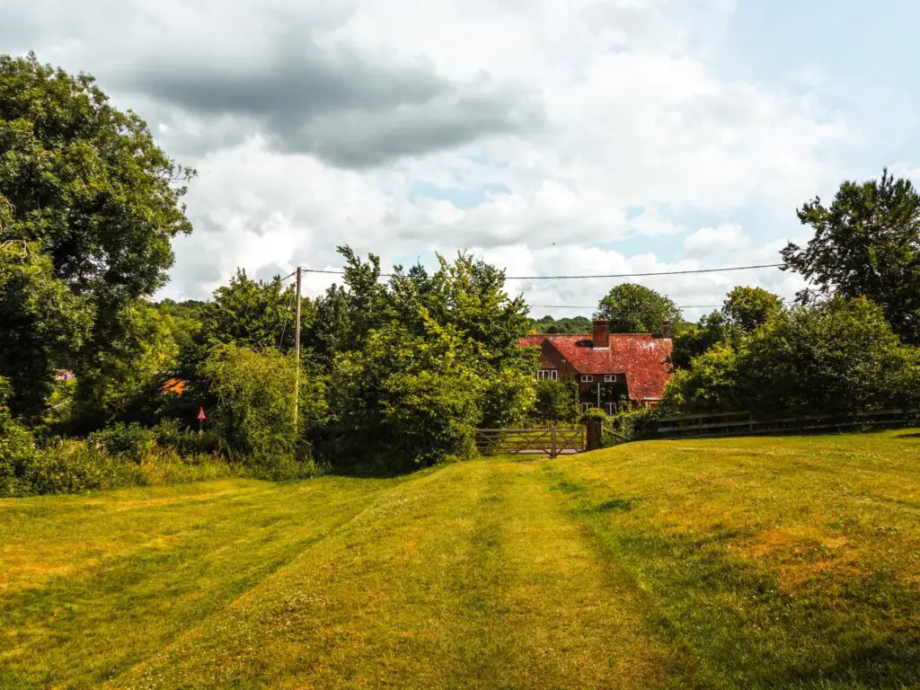 A green field with the trail running along it. There is a red house visible at the end, partially hidden by trees.