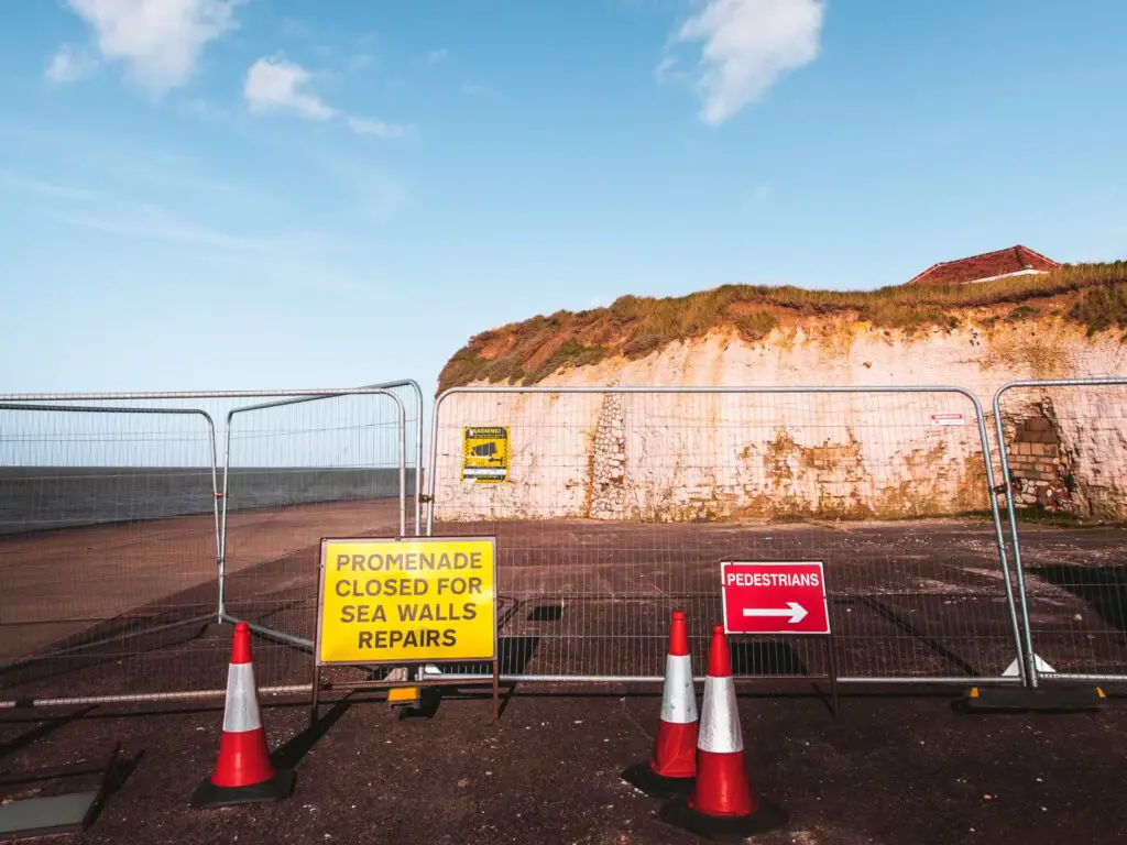 Small white cliffs with a fence in front and signs saying 'promenade closed for sea wall repairs'