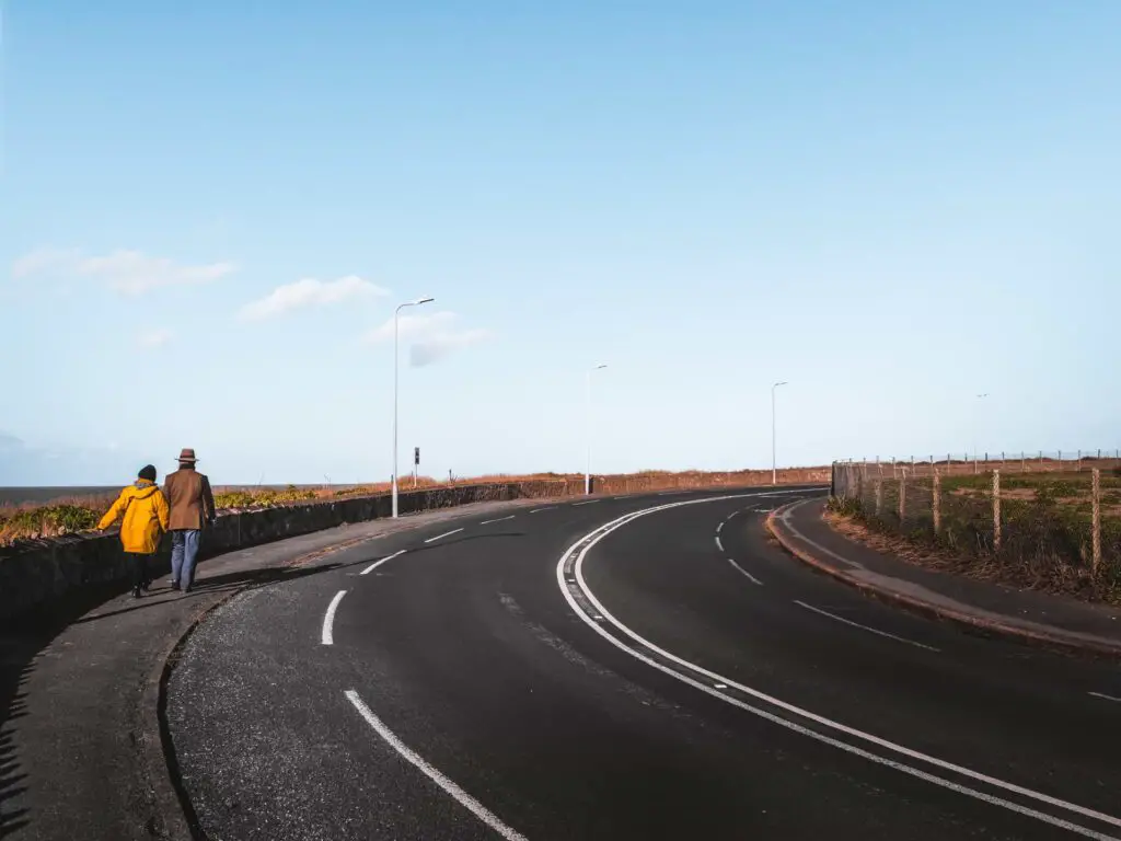 The main road as it curves around to the right. There are two people walking on the pavement on the left.