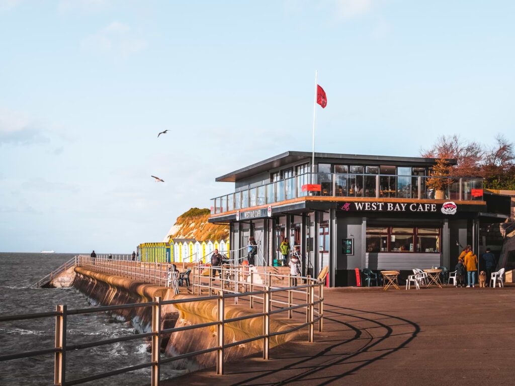 The West Bay cafe overlooking the sea. There are people walking around outside. The sky is blue.