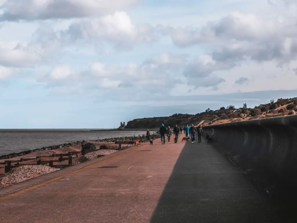 A large concrete path out of Herne Bay, with people walking their dogs, with a view of Reculver Towers in the distance.