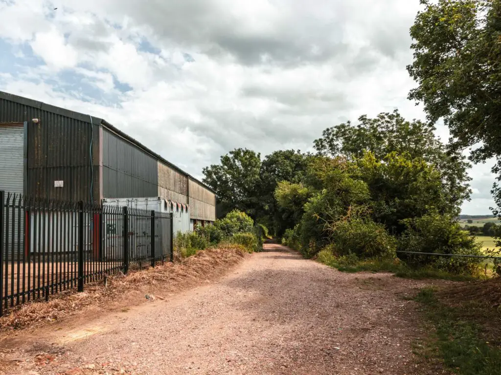 A gravel trail leading to green trees, with a farm shed and metal gate on the left. 