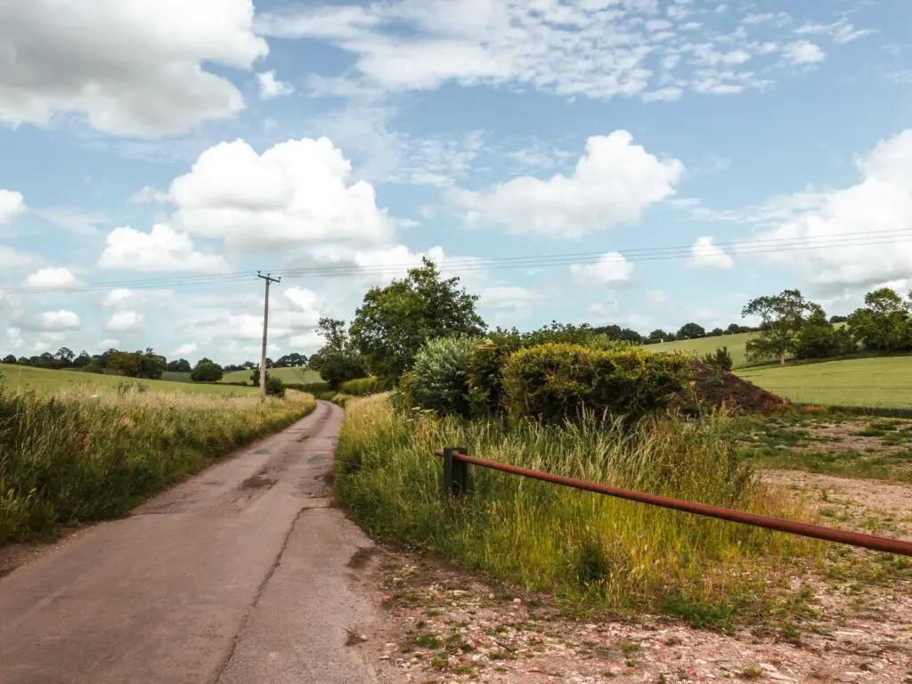 A small country lane surrounded by green fields and bushes along the circular Offley Three Springs walk near Hitchin. The sky is blue with a few white clouds.