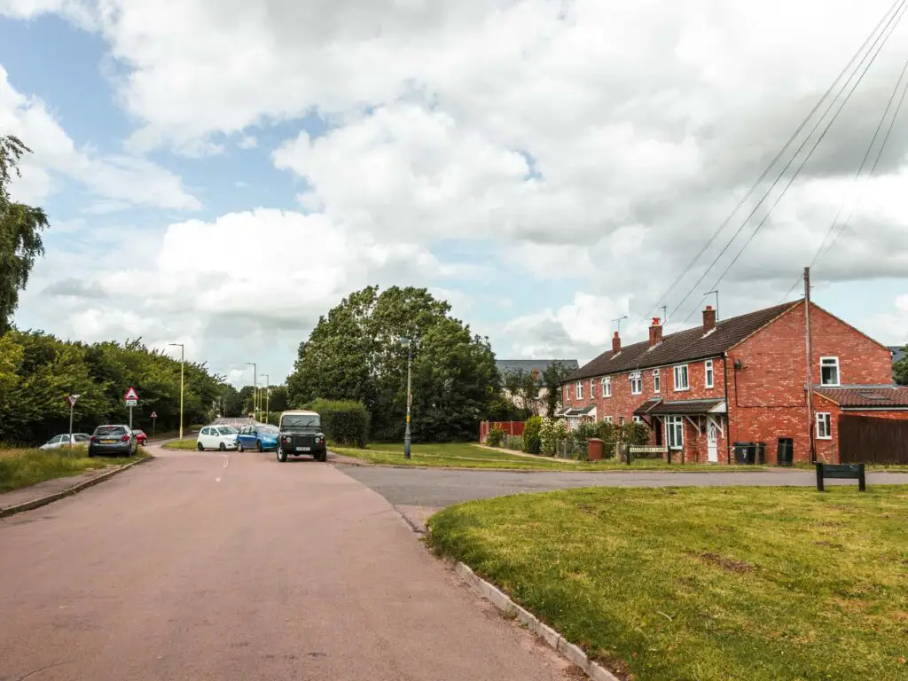The road leading to Offley village, with a side road on the right, and some pared cars up ahead. There is a red bricked building on the other side of the right side road. The sky is blue with white clouds.