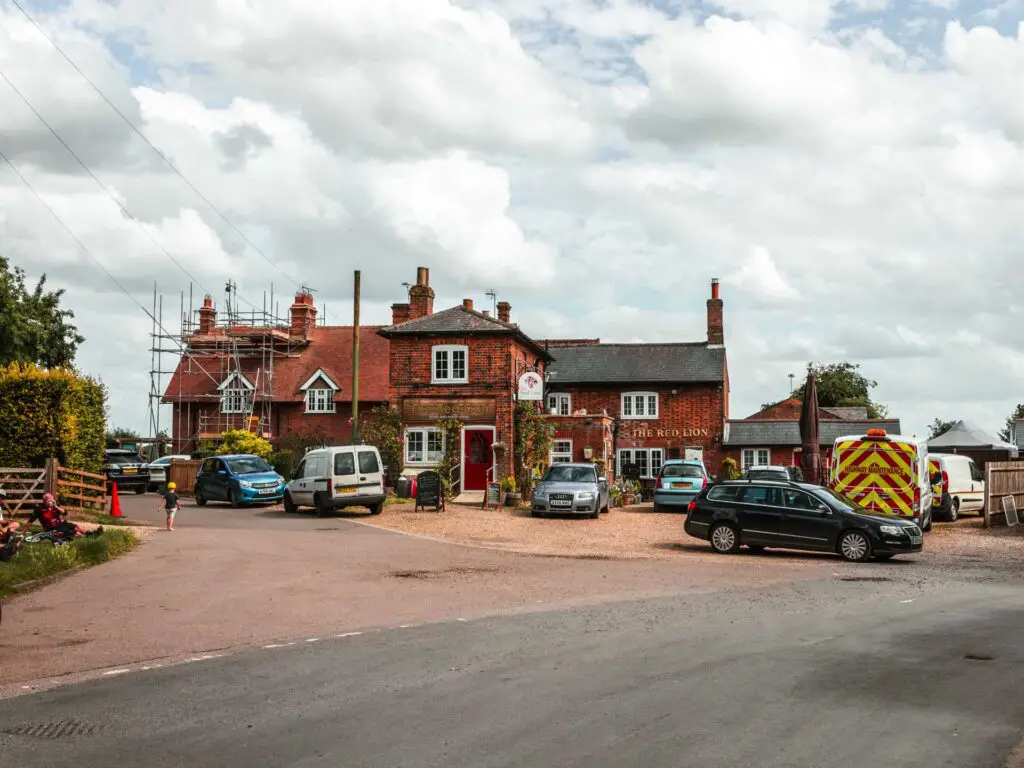 The red lion pub at the start of the Three Springs walk in Offley. There is some scaffolding on the building and a few cars parked outside. There are some people sitting on the grass on the left.