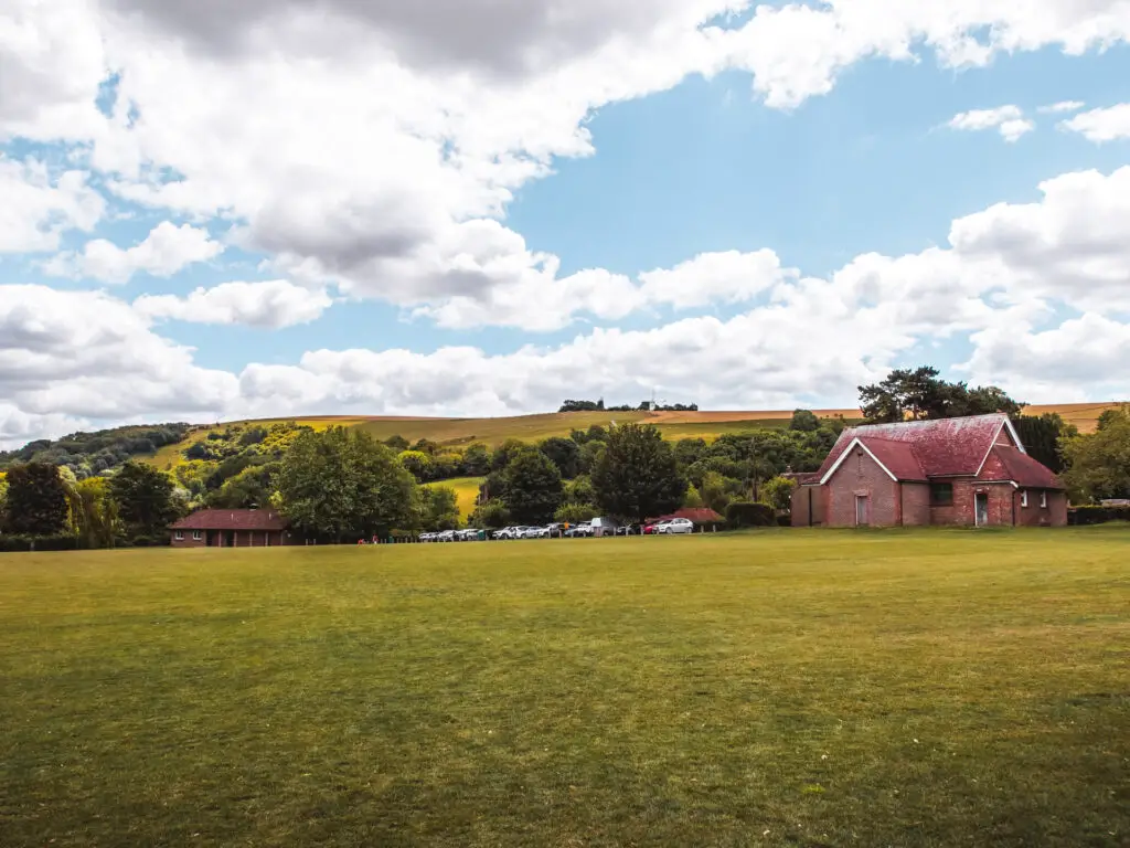 A green field with some buildings and cars on the other side and the South Downs hills behind them. 
