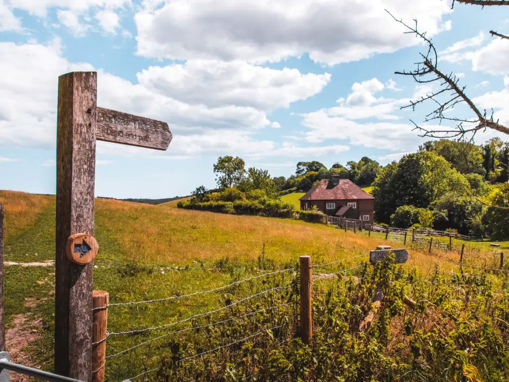 Signage next to a green feld in the South Downs, and a cute house on the other side of the field.