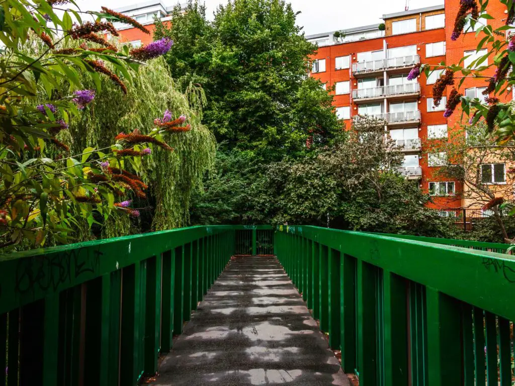 The bridge path with green railings on either side and trees and flowers lining the bridge.