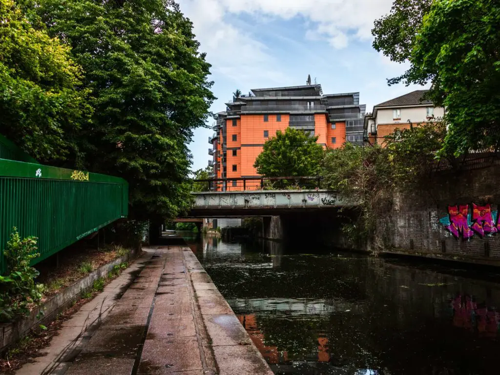 The path as it runs along the Regents Canal. There is a train bridge ahead, and graffiti on the walll on the right Sid eof the canal.