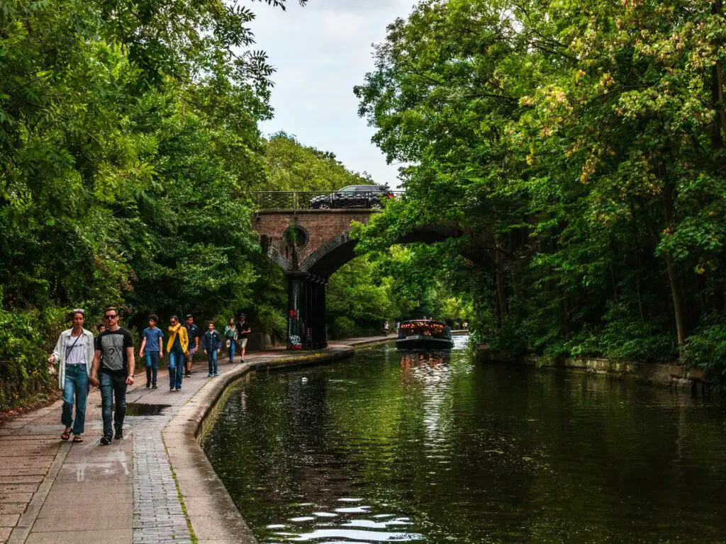 The path along the Regents Canal on the walk from Little Venice to Camden in London. There are lots of people walking on the path. There is a boat on the canal, and a bridge up ahead with a car on it.