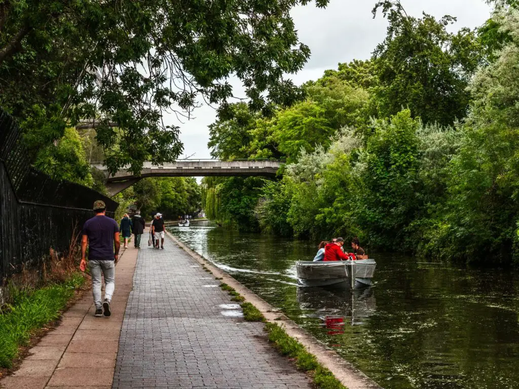 The path on the walk from Little Venice to Camden running along the Regents Canal. There are people walking on the path and there is a small boat on the water with people in it.