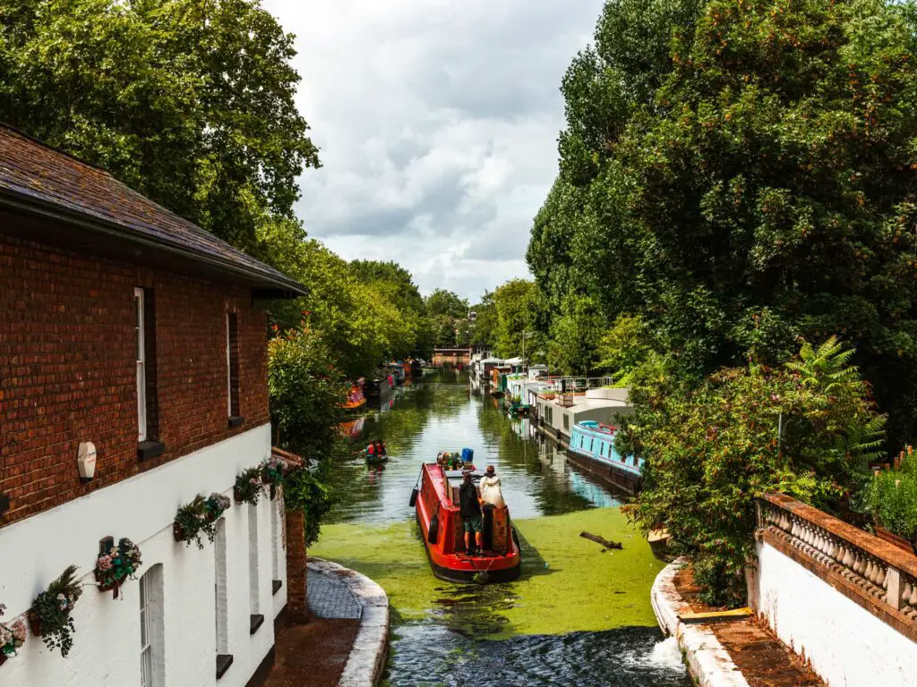 A red canal boat with people on it going along the canal at the start of the walk from Little Venice to Camden. There are canal boats and trees lining the canal. There is a white and brown brick house on the left.