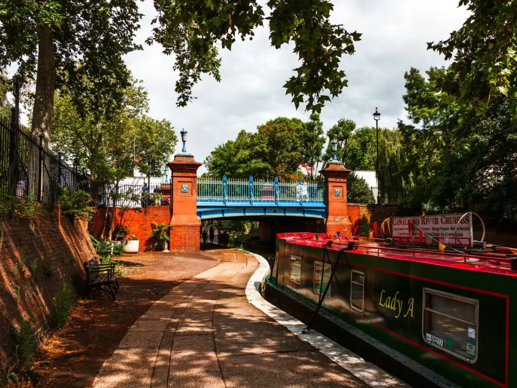 The path for the walk from Little Venice to Camden in London. There is a green and red canal boat next to the path and a blue bridge up ahead.