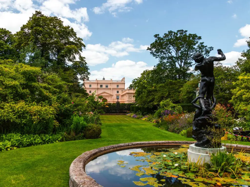 A fountain and neat green lawn with st johns lodge on the other side of the lawn behind hedges.
