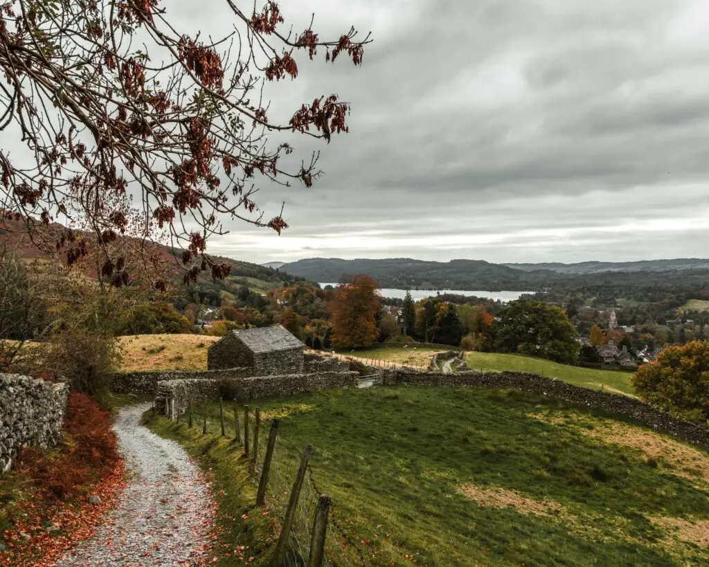 A winding gravel trail on the left on the walk up to Sweden Bridge from Ambleside. There is a green field on the right, with hills in the distance and a view of Lake Windermere. There is a small stone shed next to the trail.