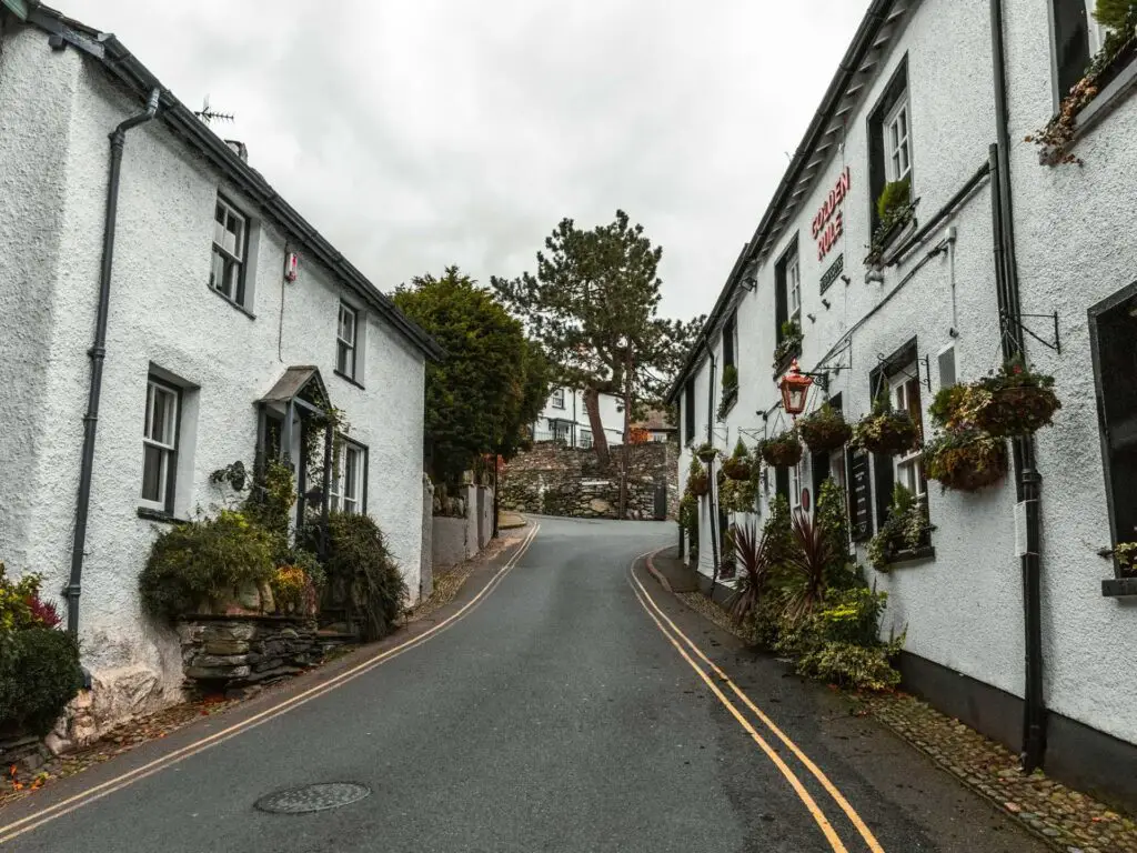 A road leading uphill in Ambleside at the start of the walk to Sweden Bridge. There are white building lining the road with plant pots hanging outside the buildings.