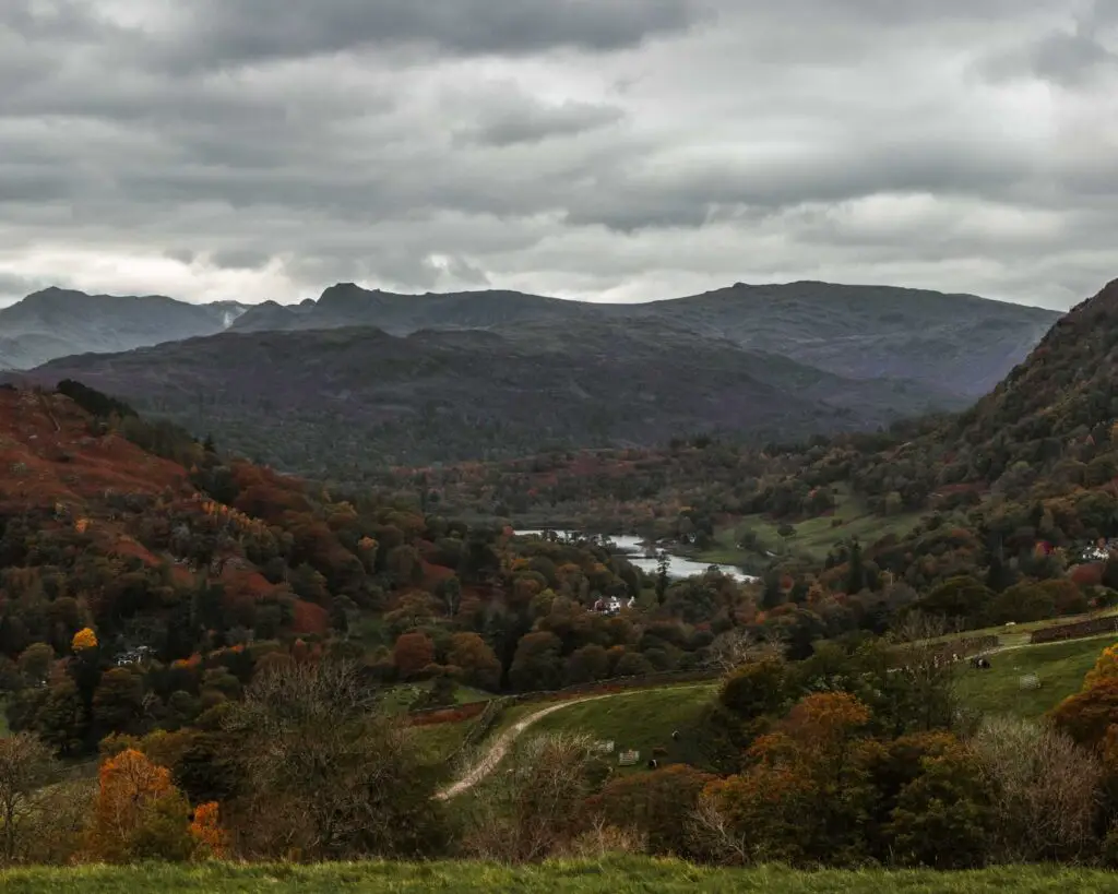 A view down to a lake in a valley on the walk to Sweden Bridge from Ambleside in the Lake District. The hills surrounding the lake a covered in trees in shades of green, orange and reds. There is cloud above the mountains in the distance.