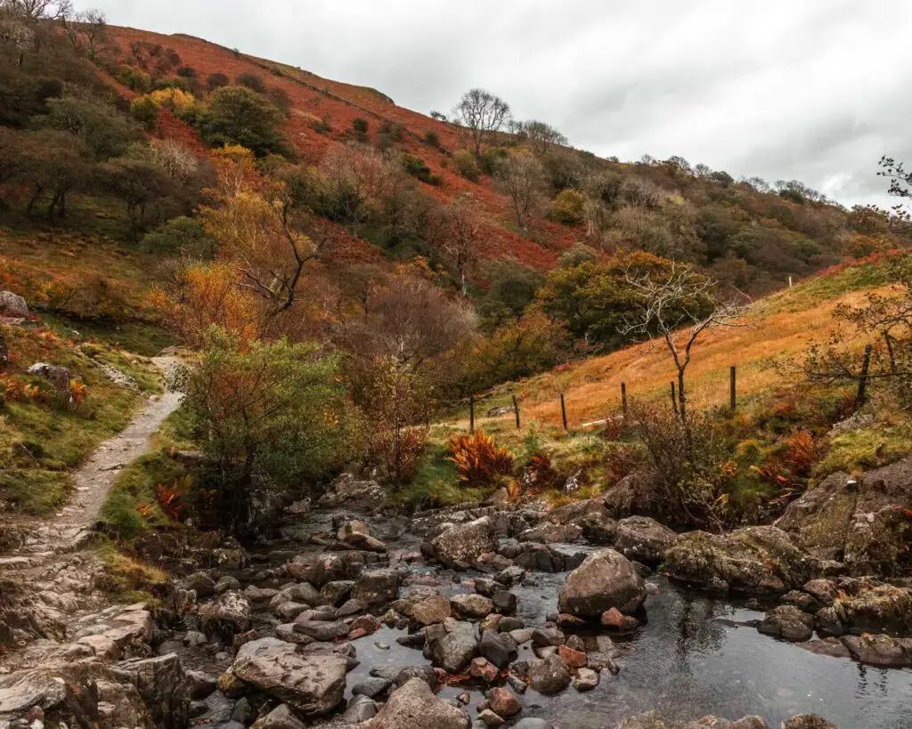 A view of the water when standing on Sweden Bridge in the Lake District. There are rocks of various sizes in the water and hills in colours of red, green and orange on  the other side.