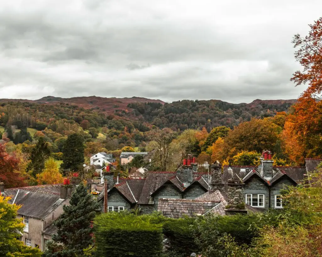 A view of rooftops of Ambleside on the uphill walk towards Sweden Bridge. The houses are surrounded by green hedges and lots of trees with leaves in Autumn shades of orange, red and green.