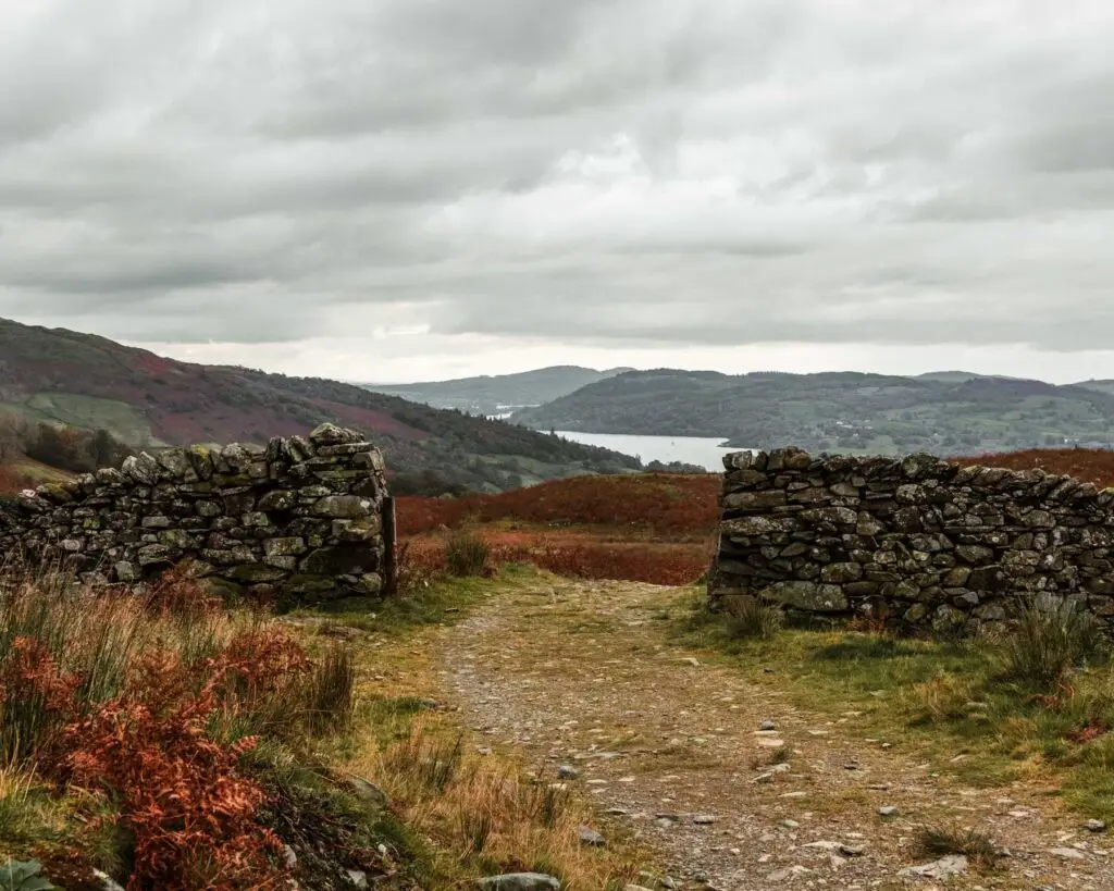 A wide gravel trail running through an opening in a stone wall with a view of lake Windermere and surrounding hills in the distance. 