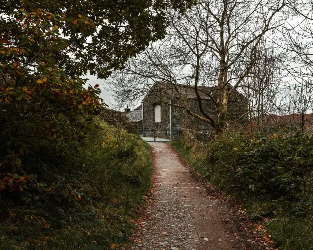 A stone farm shed ahead at the independent of the dirt, gravel trail. There are green bushes on the sides of the trail.