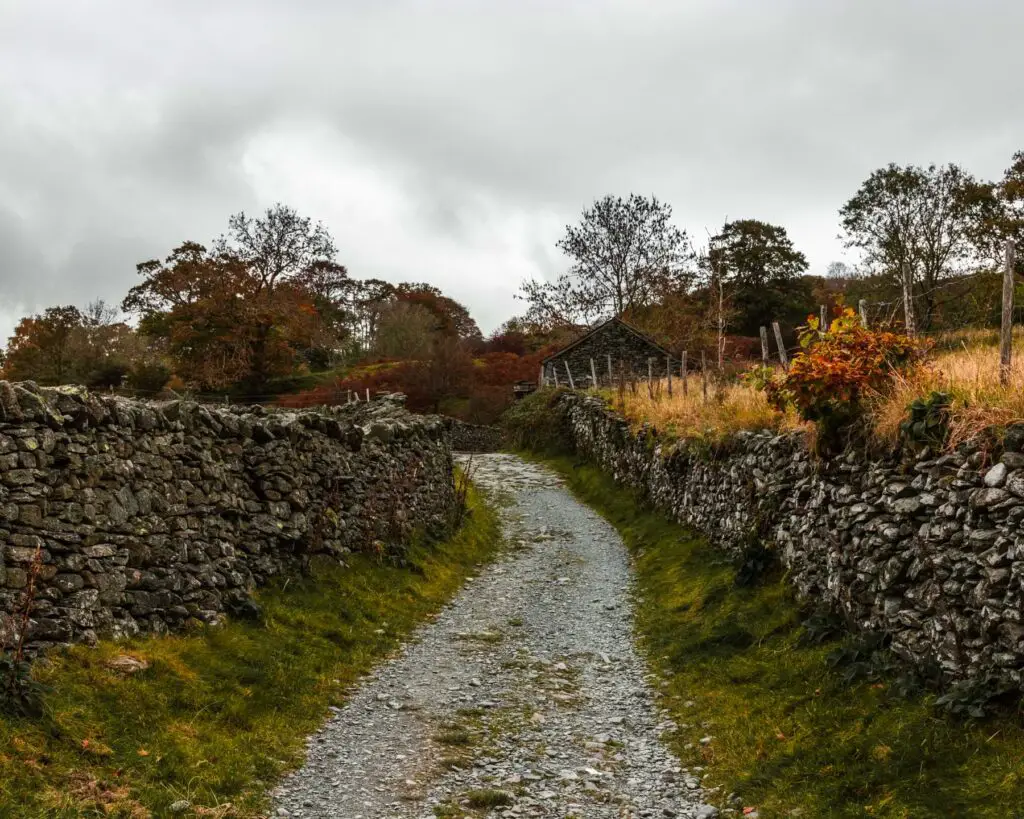 A winding gravel trail lined with green grass leading uphill, with stone walls running alongside it. 