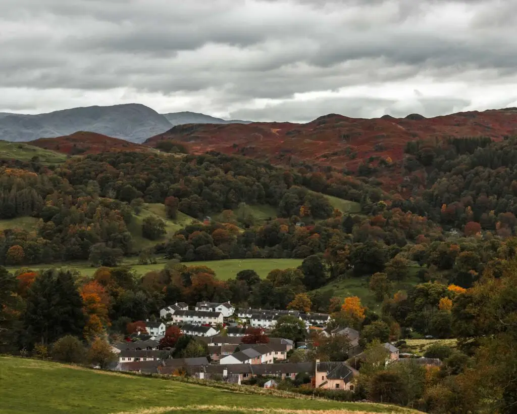 A view down to a small village on the walk up to Sweden Bridge in the Lake District. The village is surrounded by hills of green with trees and mountains in the distance. 