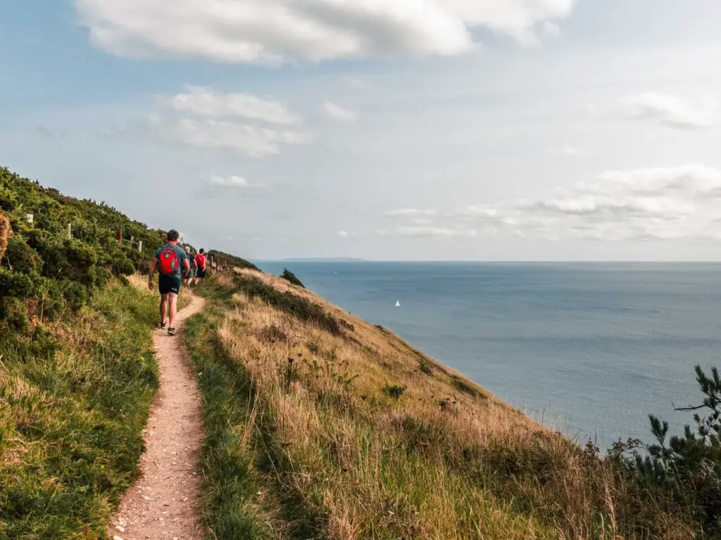 A narrow trail leading along the side of a grassy cliff on the walk to Old Harry Rocks. The blue sea is below on the right, and there are a few people walking on the trail.