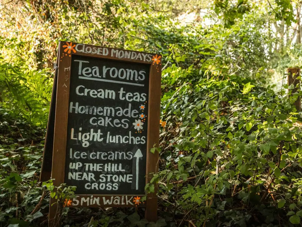 A blackboard sign nestled in the green bush. It has chalk writing for the local tea room and its menu.