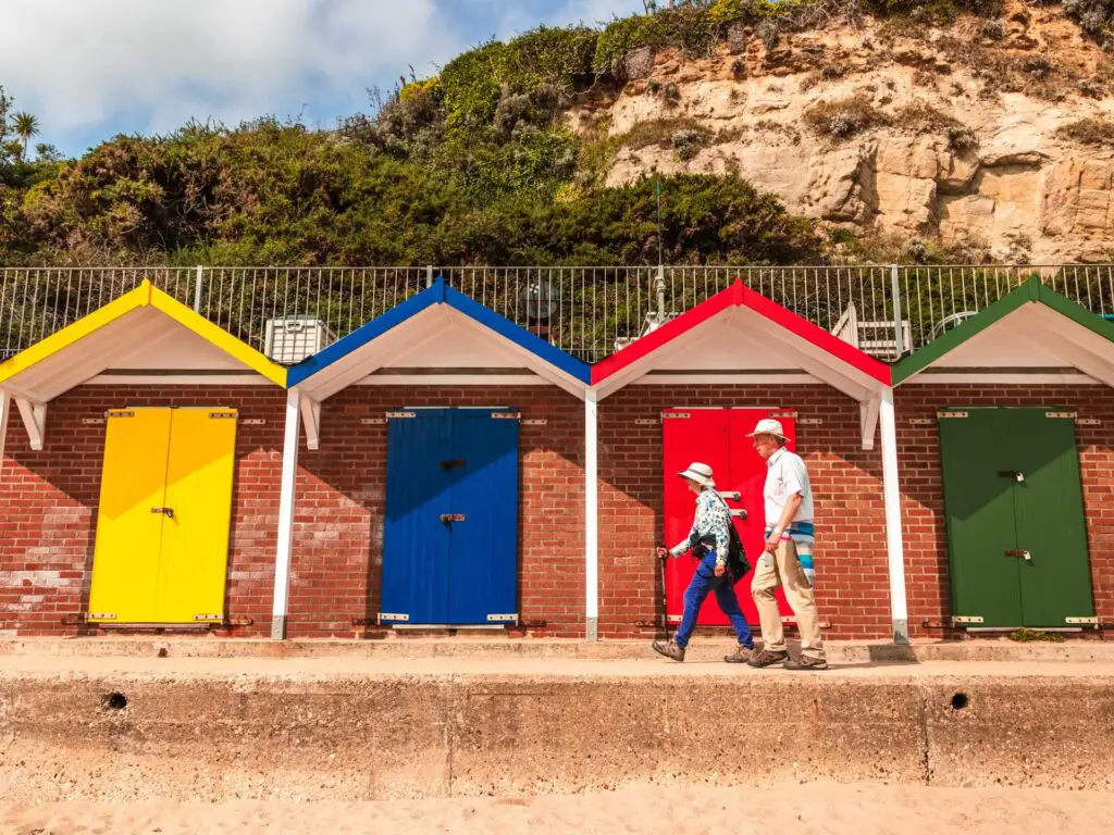 Brick beach houses with colourful doors at the start of the walk to old Harry Rocks from Swanage. There are cliffs behind the beach huts, and two people walking next to the huts.