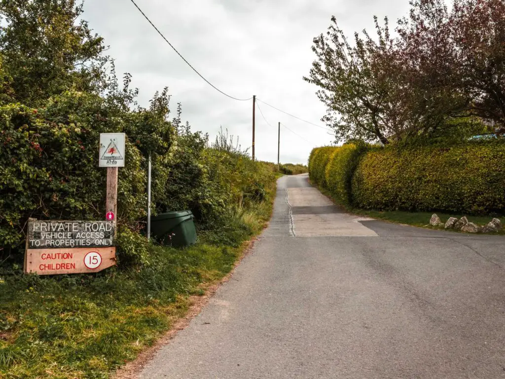 A road with a green hedge on the right and green bushes on the left. There is signage on the left marking it as a private road.