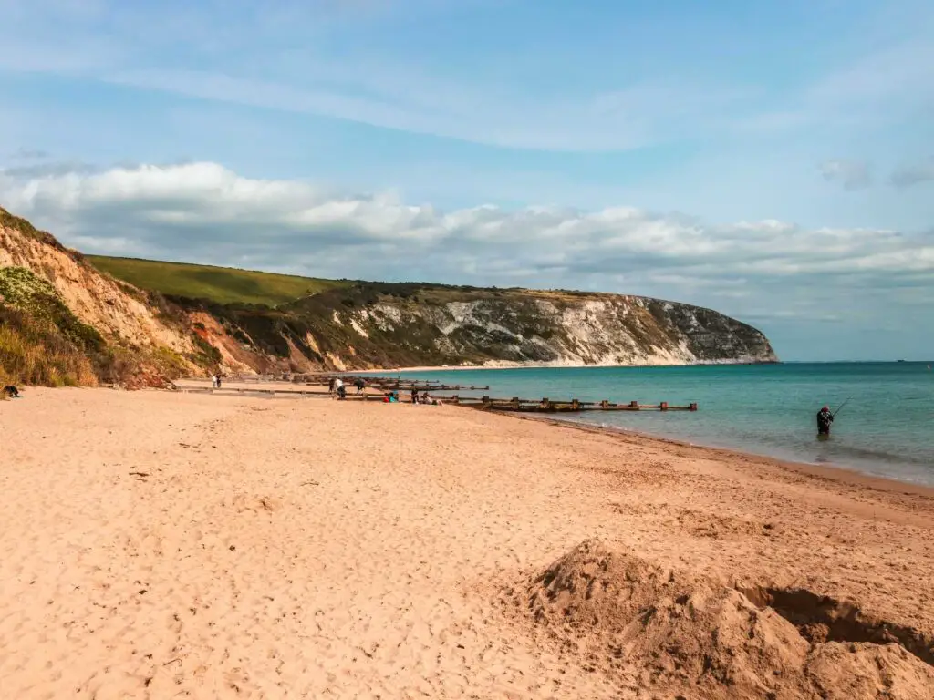 The sandy beach in Swanage with cliffs in the background leading to Old Harry rocks. The sky is blue with some white clouds. There is someone standing in the sea fishing.