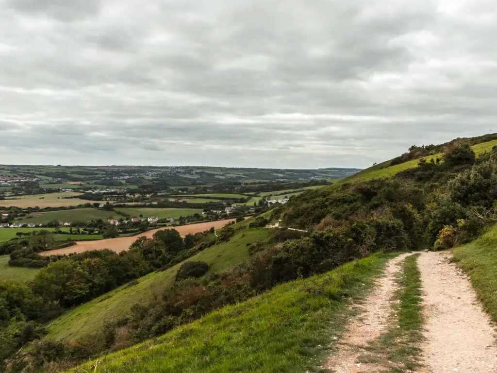 A walking trail on the right on the side of the hill with views of the green landscape in the distance. 