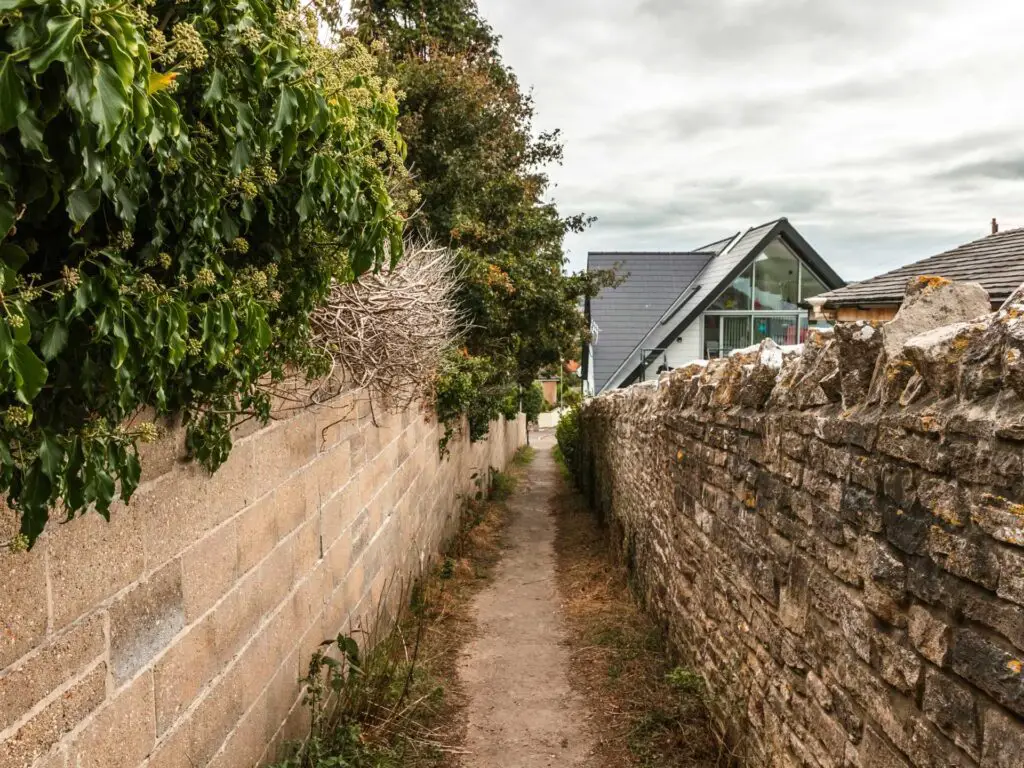 The trail leading down the back of houses. There is a stone wall on the right and brick wall on the left.