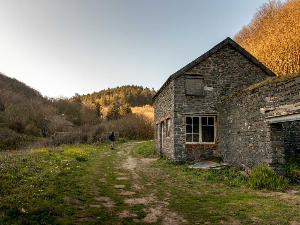 An abandoned brick building on the walk to Blackchurch Rock. There is a path and grass around the budding and trees inn the background.