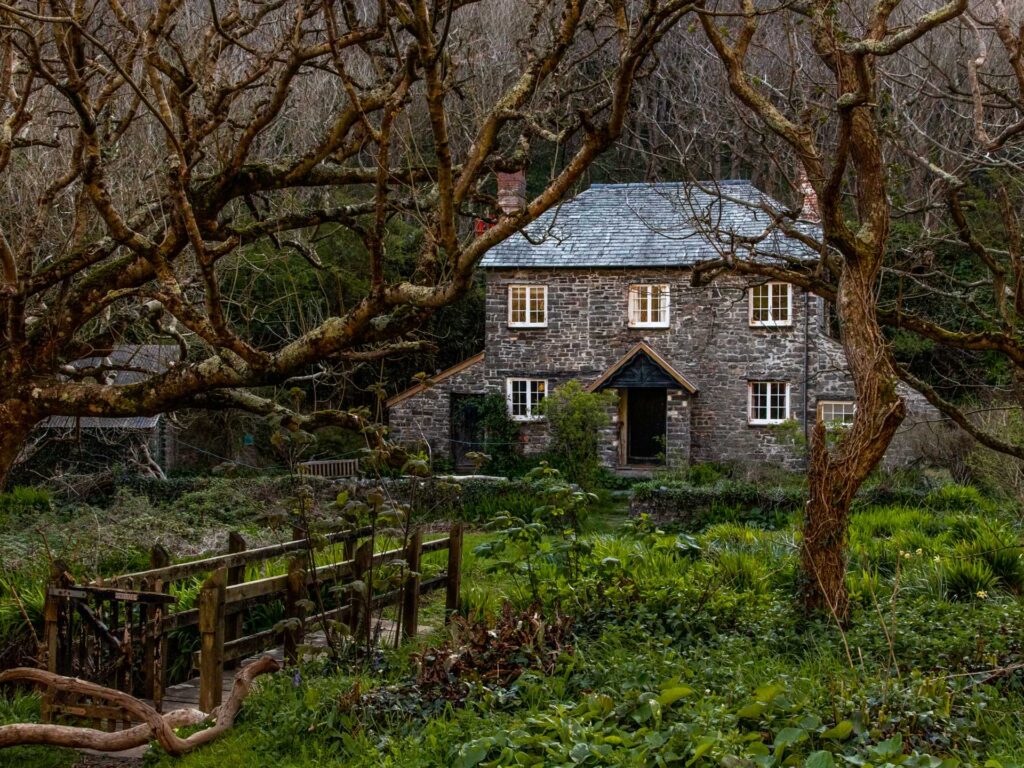 A brick cottage surrounded by trees on the walk to Blackchruch Rock and Mouthmill Beach . There is a wooden bridge leading to the cottage. 