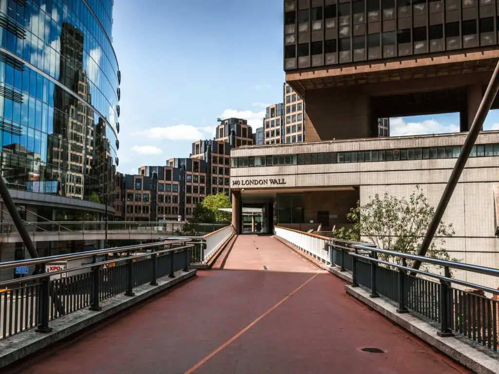 A red floored bridge walkway leading to a building ahead that says London Wall. there are tall office buildings ahead and a glass office building to the left.