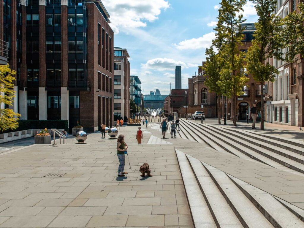 A pedestrian square in the city of London with steps and people walking around. Millennium bridge is just about visible in the distance. 