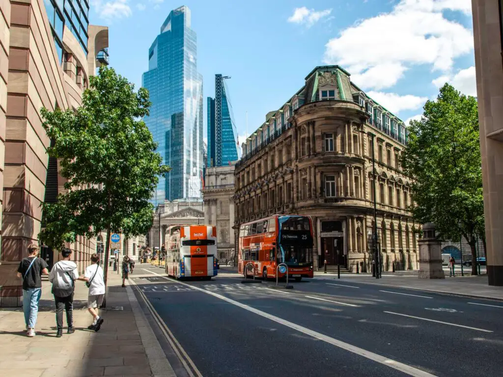 A view along the road leading towards the skyscrapers on the city of London walk. There are a few people walking on the pavement and two red buses on the road.