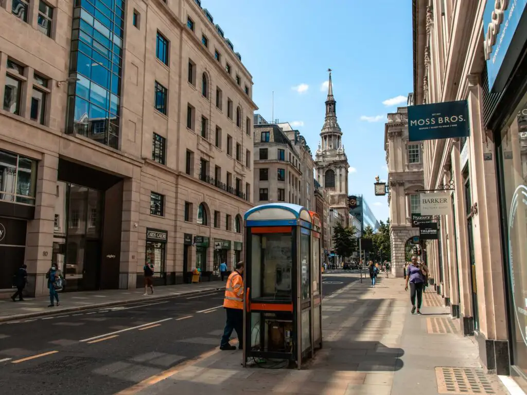 A view along a road lines with buildings in the city of London. There are a couple of telephone boxes on the pavement and a few people walking nearby.